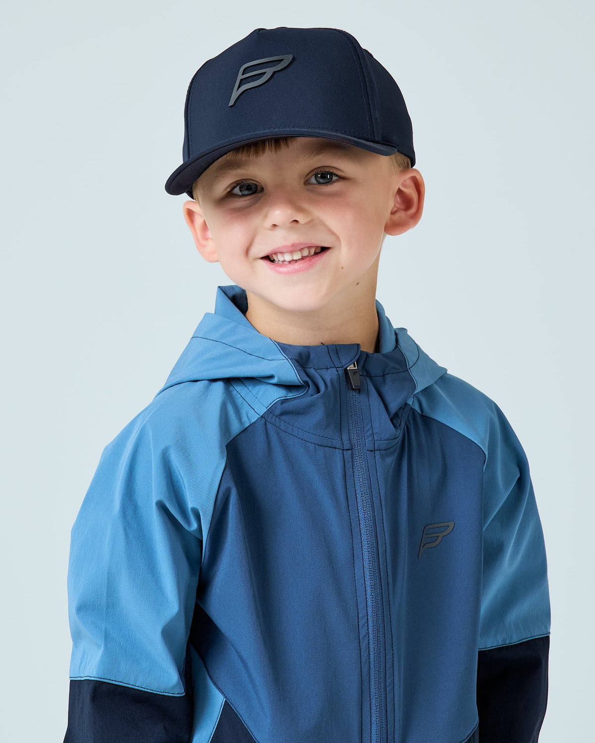 A young boy smiling while wearing a navy hooded jacket and a matching navy baseball cap with a stylized 'F' logo, set against a solid light gray background.